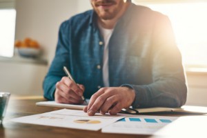 Businessman analysing statistical charts and graphs as he sits writing notes at his desk in a home office, low angle close up view of his hands